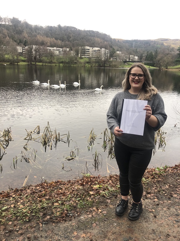 Photo of Camilla holding her thesis by the University of Stirling campus loch